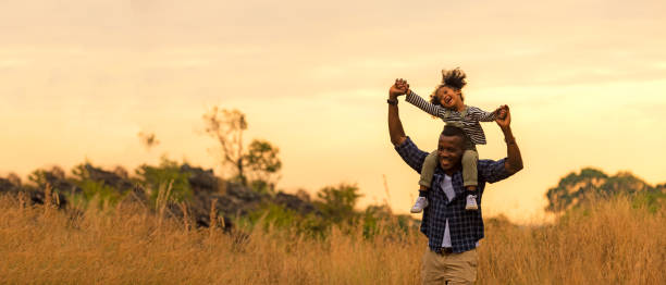 glückliche afrikanische familienkindtochter reitet den hals vater und läuft auf der wiese natur auf silhouette lichter sonnenuntergang.  reise- und familienkonzept, - ausgewogenheit stock-fotos und bilder