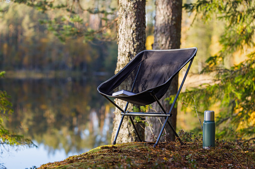 Beautiful forest lake with camping folding chair, book and vacuum insulated bottle.