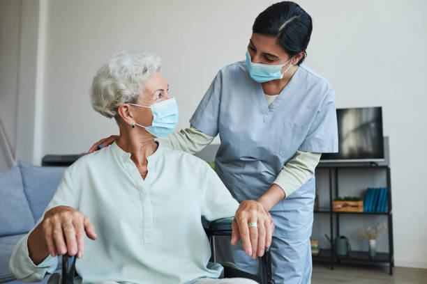 Young Nurse Helping Senior in Wheelchair Portrait of female nurse assisting senior woman in wheelchair at home or in retirement center, both wearing masks protective face mask stock pictures, royalty-free photos & images