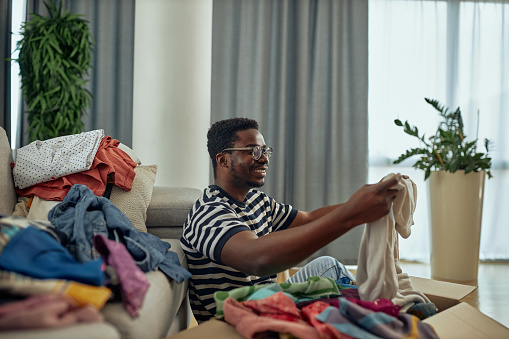 Black man sitting on the floor, leaning on sofa and sorting clothes which he putting into donation box