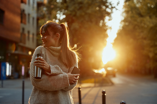 Smiling woman enjoying autumn in the city and holding phone and thermos