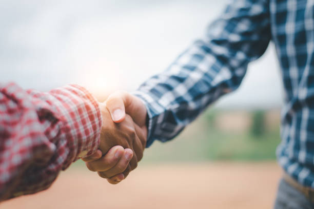 two businessmen shaking hands in field with tractor working in background. agribusiness concept - agriculture teamwork farmer people imagens e fotografias de stock