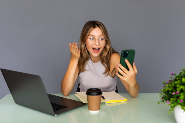 istock Smiling young female employee work on laptop in modern office use cellphone gadget browsing wireless internet. Happy woman distracted from computer job text message on smartphone device. 1351071547