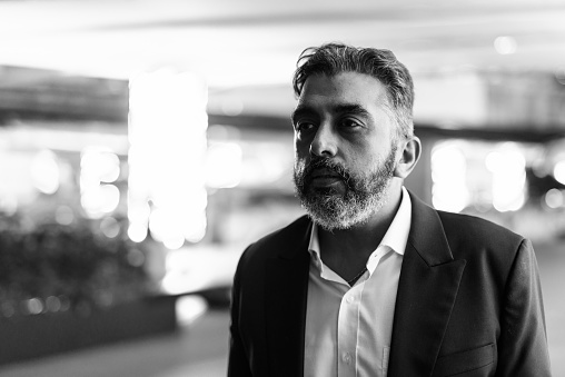 Portrait of handsome bearded Indian businessman outdoors at night shot in black and white
