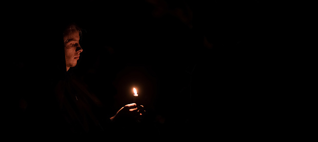 religion photography of youth monk one person only portrait in darkness with candle in hands, black panoramic background empty copy space for advertisement text, soft focus on face