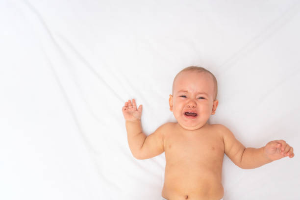 istock Hungry baby crying lying in bed on a white sheet, top view. Copyspace 1351074580