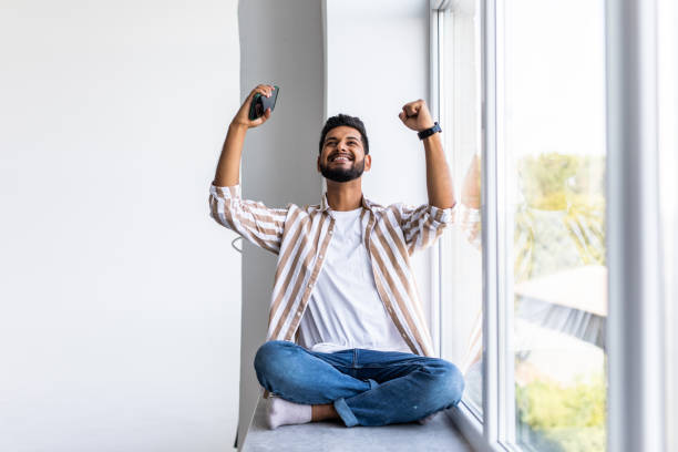 istock Indian man sitting on a windowsill in a home environment holding a phone and reading messages 1351079302