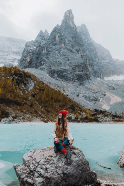 felice viaggiatrice seduta sulla roccia godendo della vista panoramica del lago di montagna ghiacciato tuqouise nelle alpi - latemar mountain range foto e immagini stock