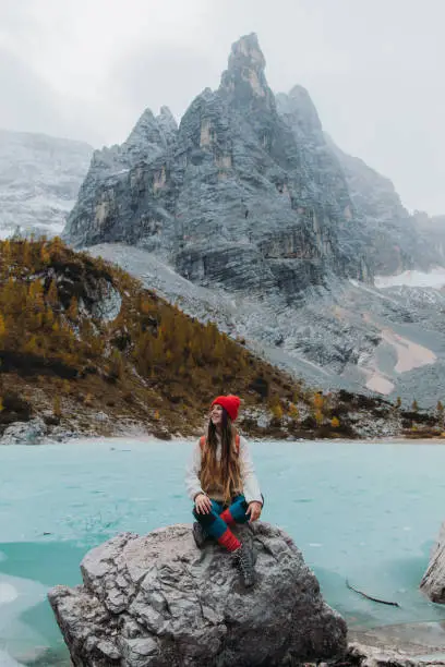 Portrait of a young woman backpacker in hat getting to the remote frozen crystal blue lake Sorapis in Dolomites Alps mountains during autumn time