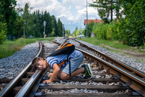 A teenager in summer clothes with a backpack sits at the crossroads of railway tracks and sadly and absently observes the surroundings. \nIt serves the arrival of the train on the tracks