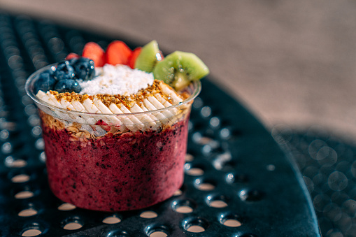 Close-Up Shot of a Healthy Acai Food Bowl with Bananas, Blueberries, Kiwi, Strawberries, Granola, Coconut, and Bee Pollen on a Picnic Table Outdoors in the Summer with Copy Space and Artistic Bokeh.