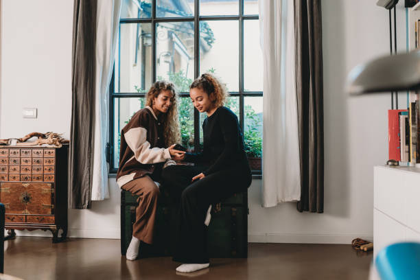 Two young adult friends sitting near a big window at home Two young adult friends sitting near a big window at home. They are spending time together. They are using a smart phone together. moroccan girl stock pictures, royalty-free photos & images