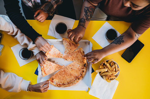 Three friends are taking slices of pizza - High angle view. A yellow table is on the background.