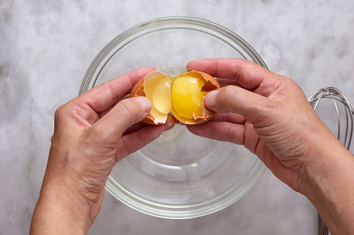 Top view of woman hand cracking raw brown egg into glass bowl on marble surface
