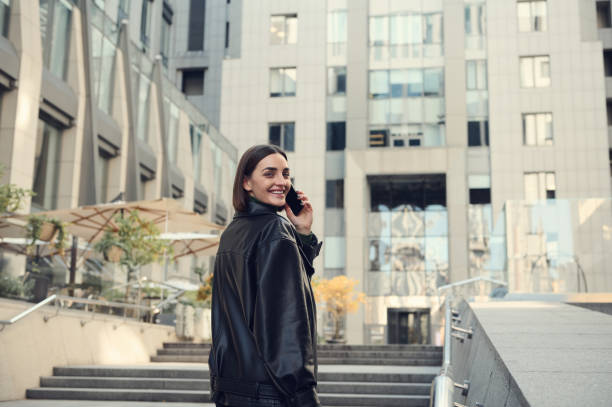 joyeuse jeune femme brune parlant sur son téléphone portable souriant sourire dentif regardant par-dessus l’épaule la caméra montant les escaliers vers un immeuble de bureaux moderne. - caméra à lépaule photos et images de collection