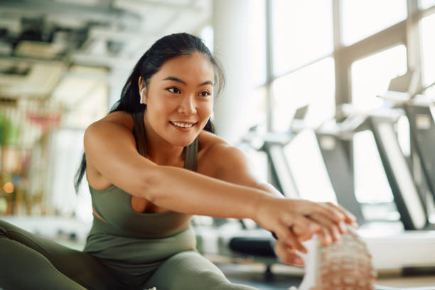 Happy Asian athlete stretching her leg while warming up for sports training in a gym Young Asian athletic woman warming up and doing stretching exercises in a gym. health club stock pictures, royalty-free photos & images