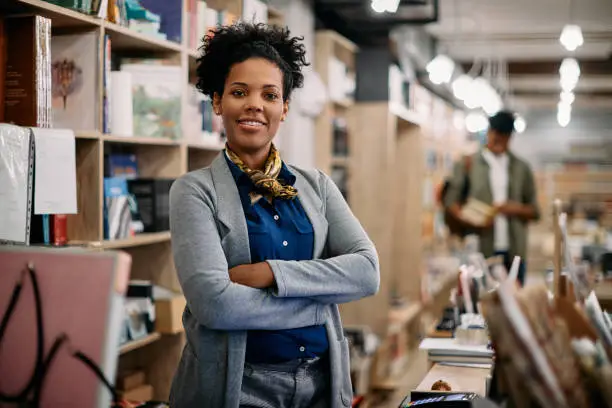 Happy African American woman standing with arms crossed while working at her book shop and looking at camera.