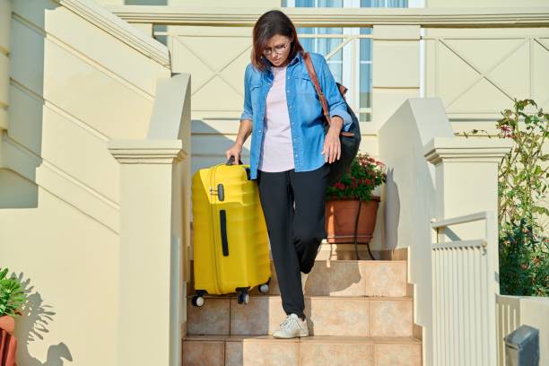 Middle-aged woman with suitcase and backpack walking down steps of house stock photo