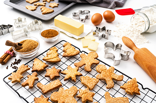 Overhead view of a white background filled with ingredients for preparing Christmas cookies. A cooling rack and a baking sheet with fresh baked cookies complete the composition. High resolution 42Mp studio digital capture taken with SONY A7rII and Zeiss Batis 40mm F2.0 CF lens