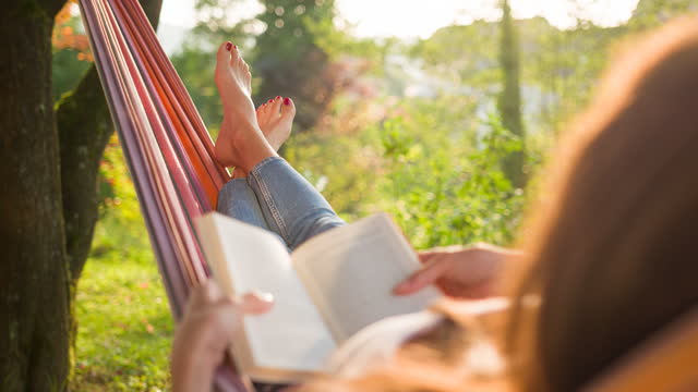 Young woman relaxing in a hammock under the trees, reading book