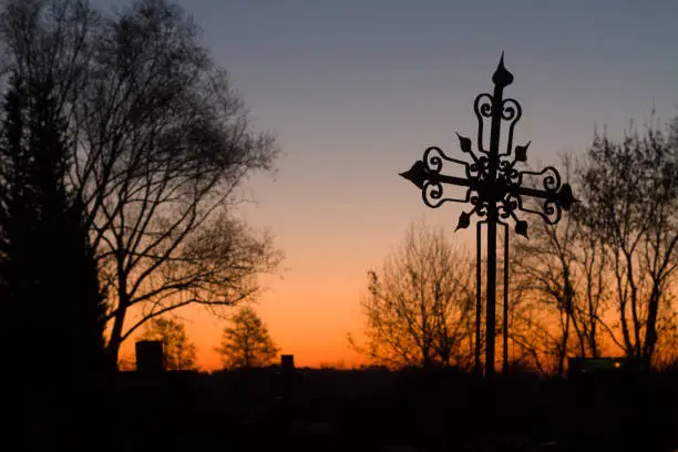 The silhouette of a metal forged cross in a cemetery before sunrise with beautiful orange-blue gradient at the sky