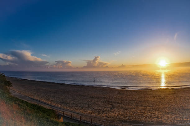 A series of four very strong and atmospheric images of late Autumn Sunrises in Sole Bay off the East Anglian coast at Southwold England in early November 2021. This fourth & final image is of a much more settled and tranquil sunrise with the sea almost flat calm, the sun as a less threatening yellow fireball and fine weather cumulus clouds scudding over the horizon, almost appearing as a tropical island sunset.