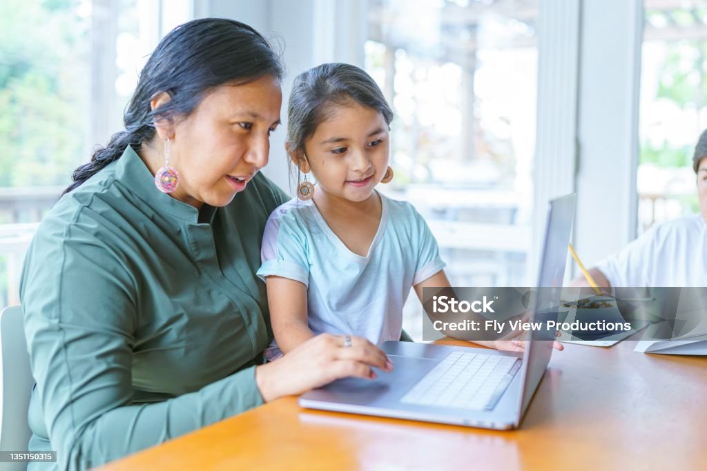 Mom working from home with homeschooled children A Native American woman sits at the dining room table and works on a laptop computer as her adolescent son works on homework next to her. The woman's elementary age daughter is sitting on her lap and is looking at the computer screen. Indigenous Peoples of the Americas Stock Photo