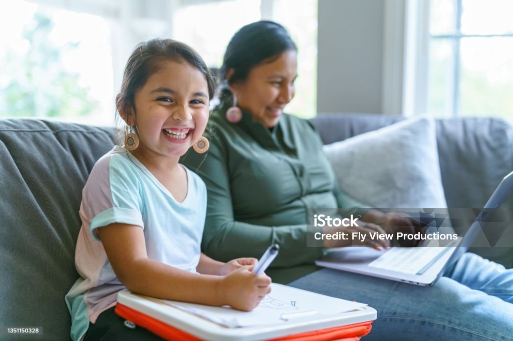 Happy Native American girl spending time with her mom Portrait of a cute elementary age Native American girl laughing and looking directly at the camera while sitting on the couch and coloring. The little girl's mom is sitting next to her and is using a laptop computer. Indigenous Peoples of the Americas Stock Photo