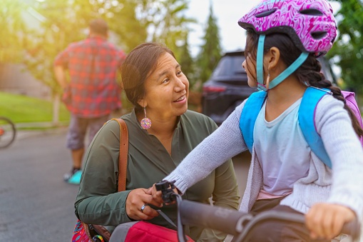 A beautiful Native American woman kneels next to her elementary age daughter who is sitting on a bike and talks with her encouragingly. The little girl is wearing a backpack and helmet and she is getting ready to leave to go to school.