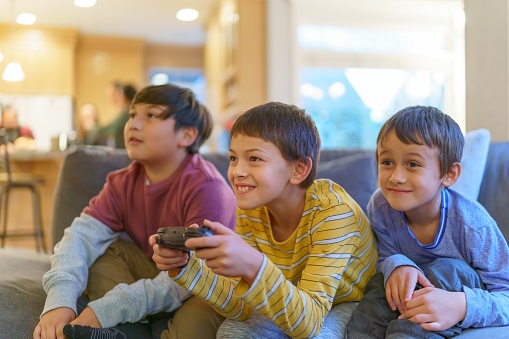 Happy Elementary age Eurasian boy plays video games with his younger brother and Native American friend at home in the living room as their parents socialize and cook dinner in the kitchen visible in the background.