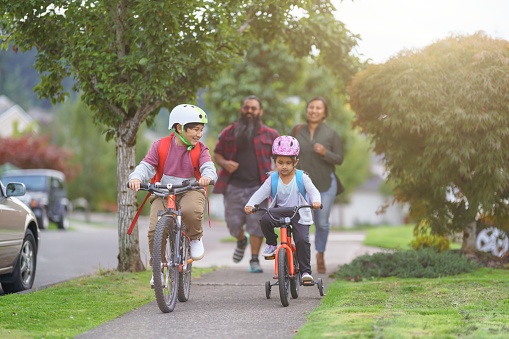 Elementary age brother and sister of Native American descent ride their bikes to school on a sidewalk through a residential neighborhood as their parents walk behind them.