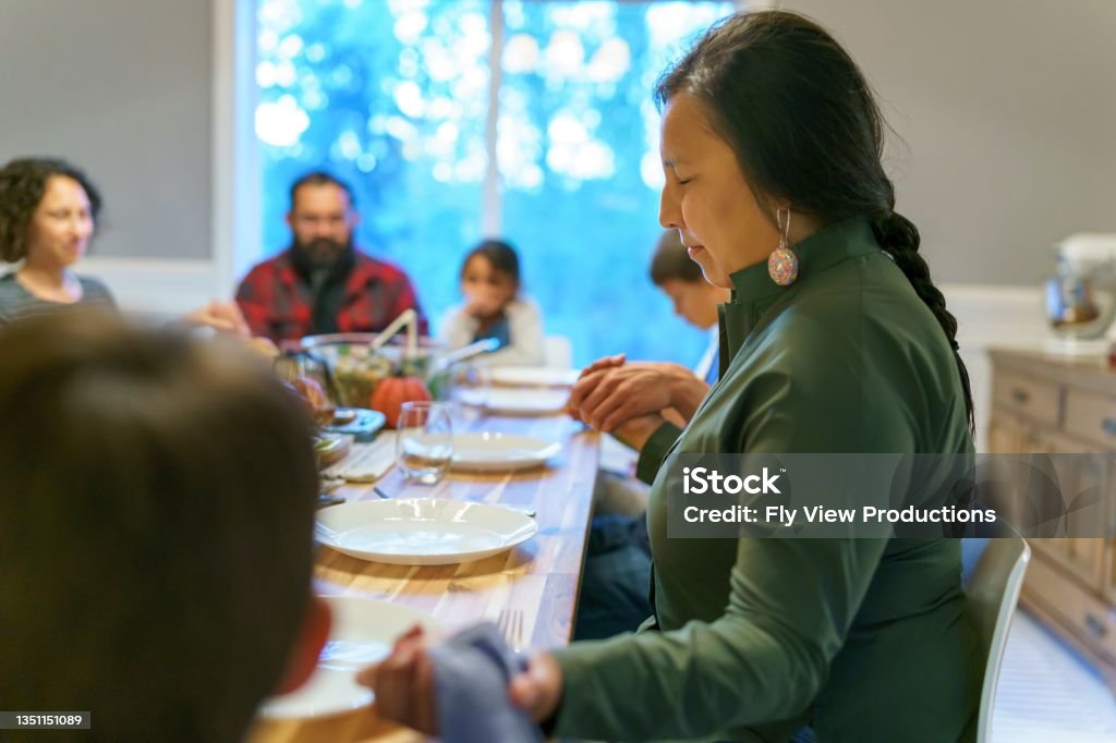 Friends and family holding hands and praying before holiday dinner Multi-ethnic group of adults and children hold hands and give thanks with prayer while sitting around dinner table before holiday meal with friends and extended family. Indigenous Peoples of the Americas Stock Photo