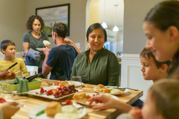 Family eating together at holiday table Multi-ethnic group of adults and children of Eurasian, Native American and Caucasian descent sitting around a dining room table filled with food for a traditional winter holiday dinner serving themselves and eating while celebrating with family and friends. indian dinner stock pictures, royalty-free photos & images
