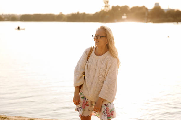 This stock photo shows a young blonde woman walking on the beach in a dress and white sweater with flowers on a sunny day in spring