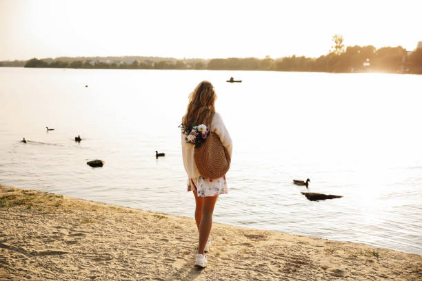 Girl On A Beach Walk This stock photo shows a back young blonde woman walking on the beach in a dress and sweater with straw bag and flowers on a sunset. straw bag stock pictures, royalty-free photos & images