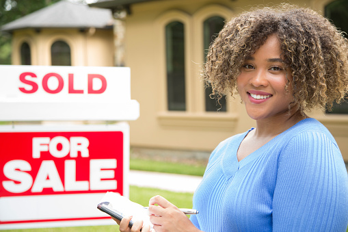 Lovely young adult Real Estate Agent standing beside her for sale sign in front yard of home.  She holds a portfolio and wears a blue top and jeans.  Sold Sign.