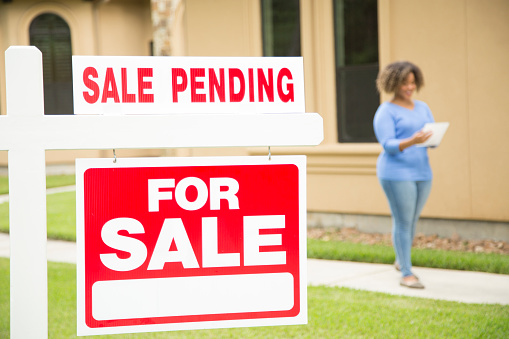 Lovely young adult Real Estate Agent standing beside her for sale sign in front yard of home.  She holds a portfolio and wears a blue top and jeans.  Sale Pending Sign.