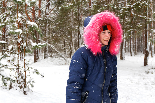 Portrait of smiling happy girl in pink warm hood walking outside on nature winter snowy forest background. Pretty child outdoor, cold weather. Caucasian kid. Winter joy.