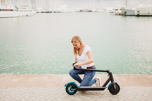 Young blonde woman folding or unfolding up her motorised scooter. Ecological mean of locomotion vehicles and green eco energy with zero emission concept. Creative color editing with added grain. Selective focus. Part of a series.