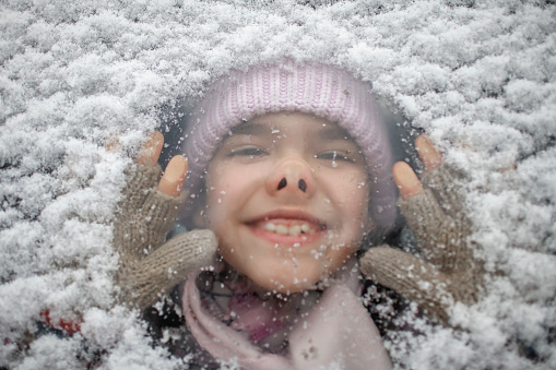 Funny portrait of little girl in warm cap through the snowy car glass, smiling and laughing, during family road trip to forest on snowing day