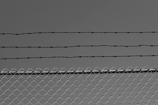 A black and white shot of fences and barbed wires in foreground and brick wall in the background, representing a jail or a penitentiary building - stock photography
