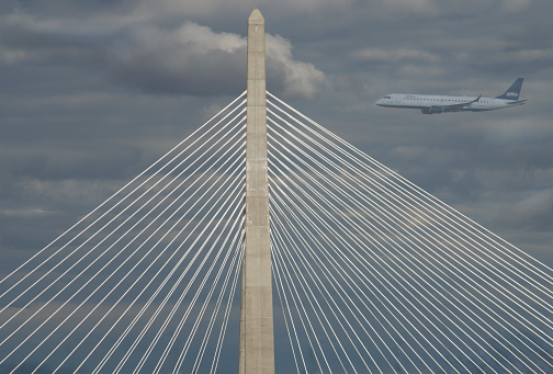 A cloudy sky contrasts with the distinctive steel cables of the Leonard P. Zakim Bunker Hill Memorial Bridge, which spans the Charles River in Boston.  And, of course, there is always a plane heading to or from Logan Airport on the edge of the city.
