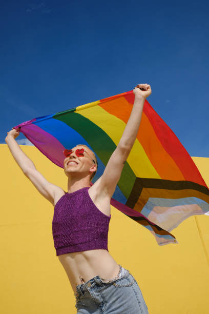 istock Low angle view of a young happy non-binary person holding and waving a rainbow flag over head. 1351218924