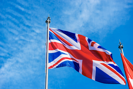 Closeup of a national UK flag with flagpole, blowing in the wind on a blue sky with clouds and copy space. Union Jack Flag.