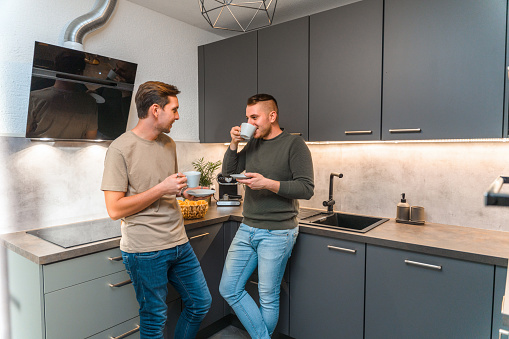 Young adult gay couple drinking coffee. Standing in a kitchen spending quality time talking to each other.