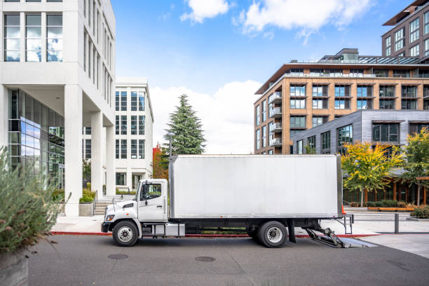 Profile of day cab medium size semi truck with long box trailer unloaded delivered goods to new multi-level apartments in unban city area White middle class relocation rig day cab semi truck tractor with long spacious box trailer standing on the urban city street with multilevel apartment and office buildings unloading delivered goods chassis photos stock pictures, royalty-free photos & images