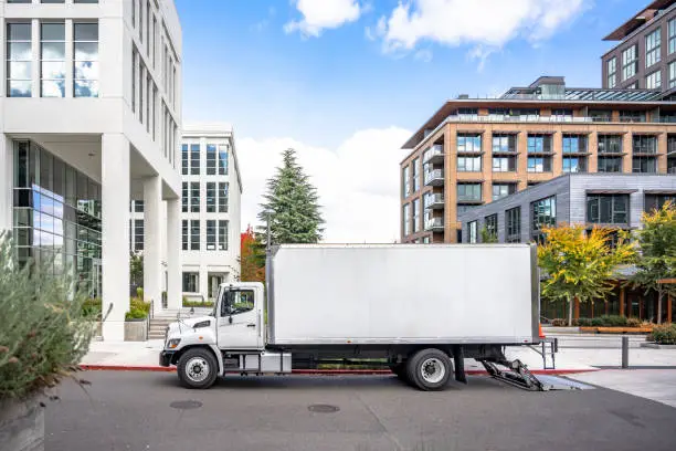 Photo of Profile of day cab medium size semi truck with long box trailer unloaded delivered goods to new multi-level apartments in unban city area