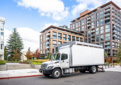 White middle class relocation rig day cab semi truck tractor with long spacious box trailer standing on the urban city street with multilevel apartment and office buildings unloading delivered goods\