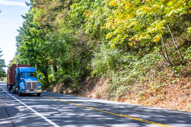 istock Popular blue big rig semi truck transporting container on flat bed semi trailer moving on the mountain road with forest on the side 1351230052
