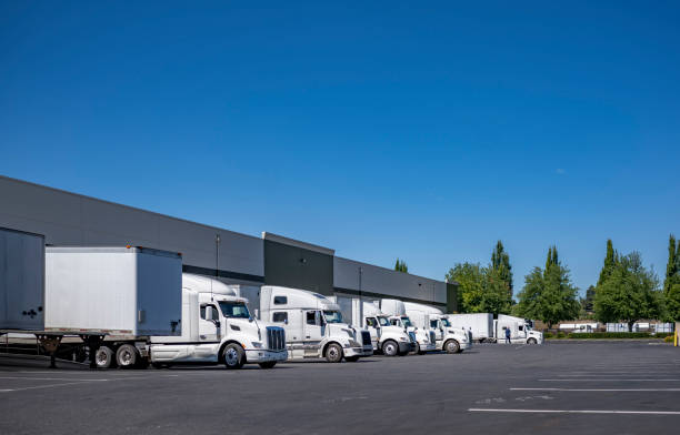 istock Row of the big rig semi trucks with semi trailer standing at warehouse dock loading cargo for the next freight 1351231533